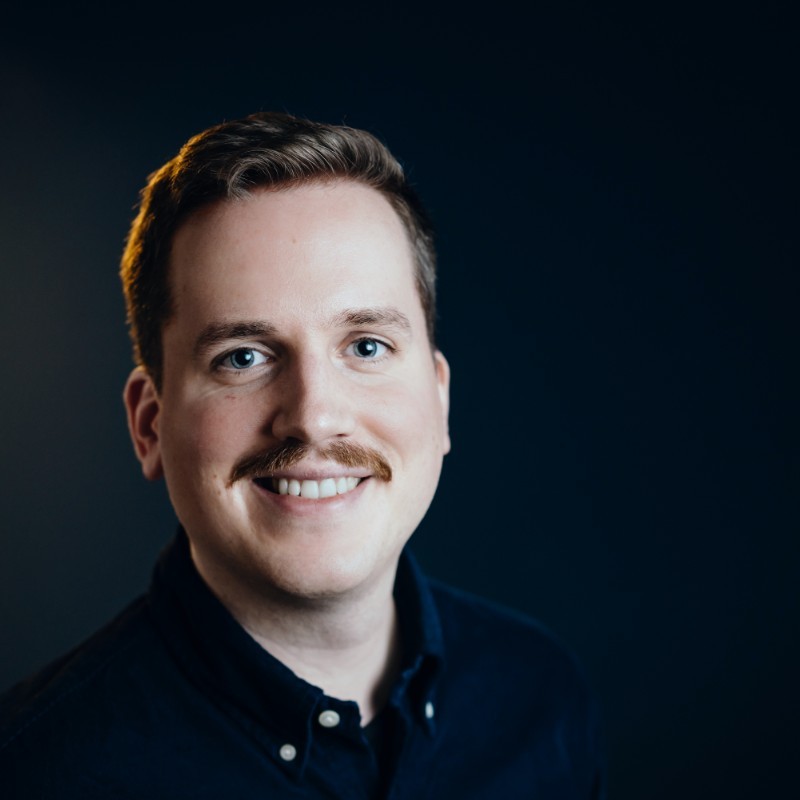 Smiling man with mustache, dark shirt, dark background.