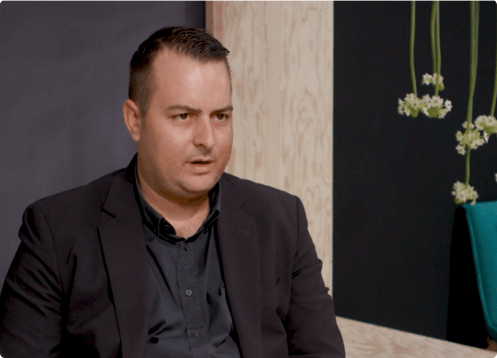 Man in black suit sitting with a neutral expression.