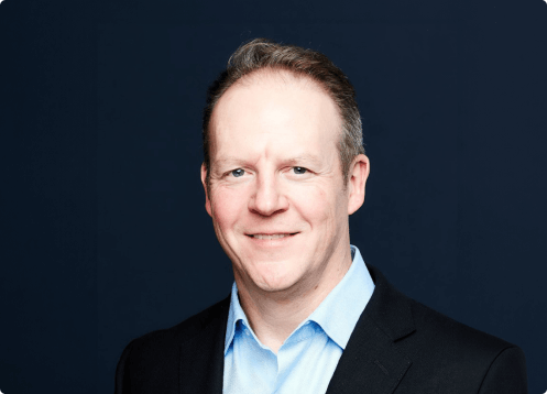 Man smiling in a suit with dark background.